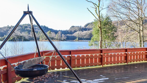 Eine Holzterrasse mit einem hängenden Grill, mit Blick auf einen See und bewaldete Hügel.