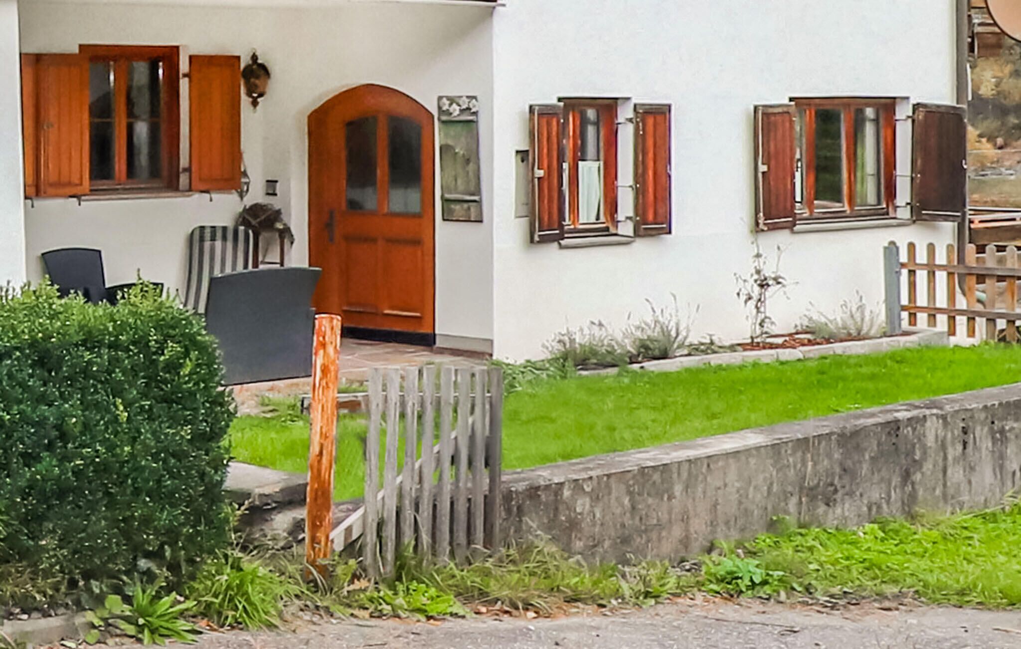 A two-story building with a white facade, wooden shutters on the windows, and a front door with an arched design. There is a small garden area with a wooden fence and a bench.