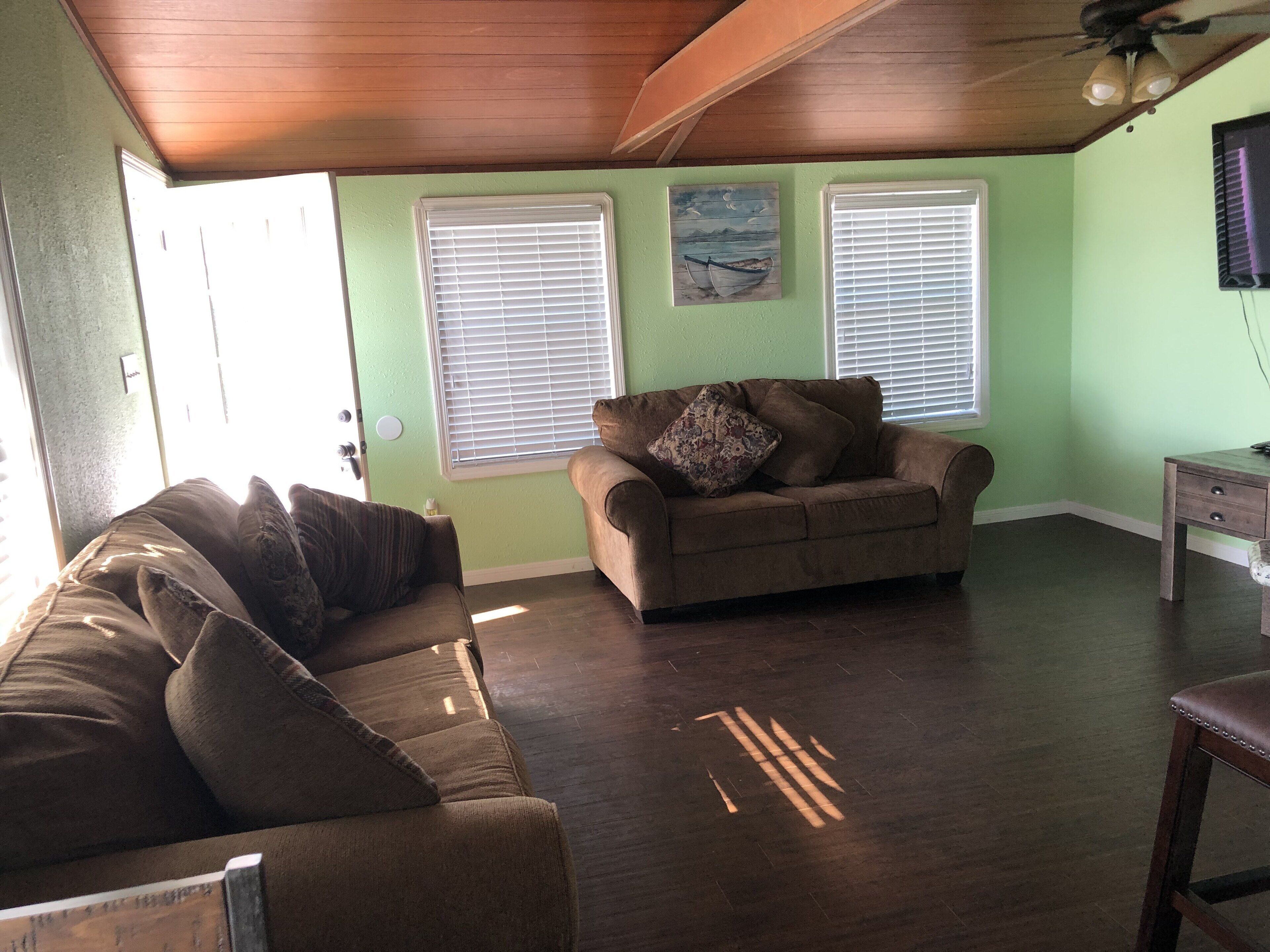 A living room with a brown couch, a wooden ceiling, and two windows with blinds.
