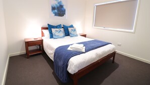 Una cama bien hecha con una colcha y almohadas azules, una mesita de noche con lámpara y un ventanal con persianas.