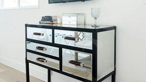 A modern living room with a glass-fronted black console table, a mounted flat-screen TV, and a large window providing natural light.