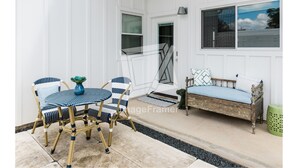 A patio with a round table and two chairs, a wooden bench with cushions, and a green trash bin.