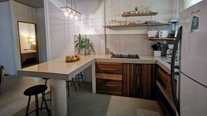 A modern kitchen with a central island, wooden cabinets, and a built-in stove. There are bar stools, a hanging light fixture, and a shelf with kitchenware. - Casa mindanao apartments, AC+WIFI + swimming pool. (Limon)