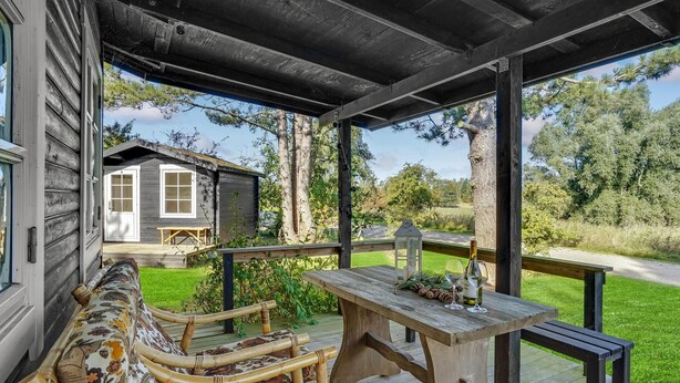 Eine Holzterrasse mit Bank und Tisch, im Hintergrund eine kleine Hütte und Blick auf die umliegende Grünfläche.