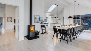 Een moderne keuken met een houtkachel, een eethoek met een lange tafel en stoelen, en uitzicht op een blauwe lucht door grote ramen.