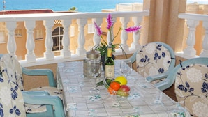 Un balcon avec une table dressée pour deux personnes, agrémentée d’une nappe à motifs floraux, de deux chaises, d’une bouteille de vin, d’un verre et d’un saladier de fruits.