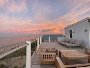 A wooden deck with lounge chairs and tables overlooking a beach, with a building in the background. - Stunning, light-filled beachfront sanctuary with full views of Delaware Bay. (Prime Hook Beach)