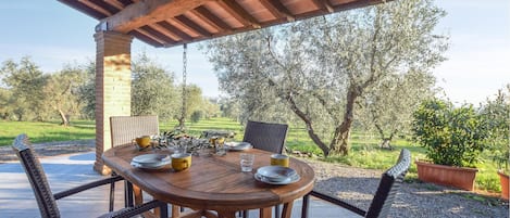 En uteplats med ett träbord dukat för middag, omgiven av flätade stolar, under en träpergola.