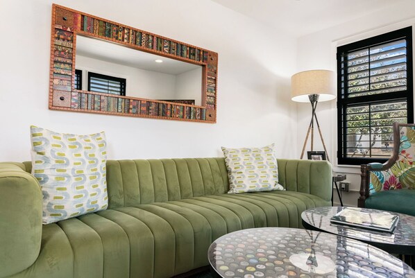 Un sofá verde con almohadones estampados, una mesa redonda de vidrio, una lámpara de pie y una estantería con libros.