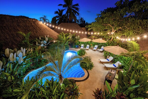 A swimming pool surrounded by lounge chairs and umbrellas, with string lights and tropical plants.