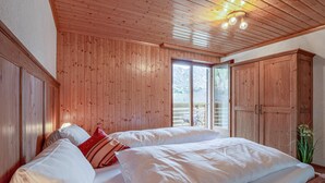 Un dormitorio con paredes de paneles de madera, una cama grande con ropa de cama blanca, un almohátero rojo y vistas a un balcón con una silla.