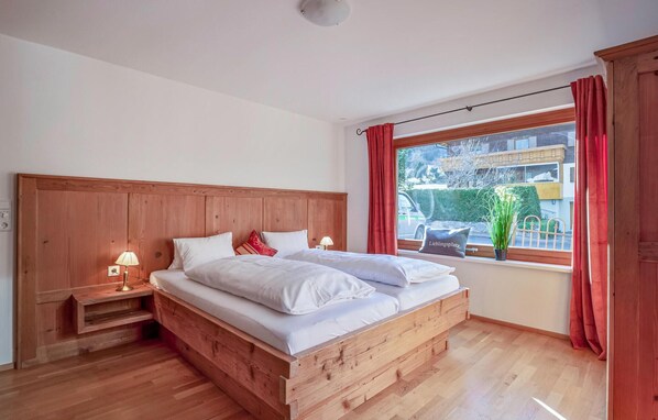 A wooden bed with white bedding, a nightstand with a lamp, and a window with red curtains. - großes Ferienhaus St. Gallenk. (St. Gallenkirch)