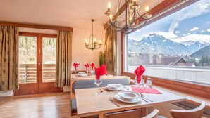 Comedor con una mesa dispuesta para cuatro, una araña y vistas a montañas a través de amplios ventanales.