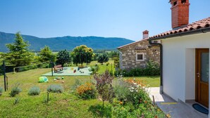 A stone house with a red-tiled roof and a chimney, a small garden with flowers and a play area, and a mountainous backdrop. - Awesome home in Krsan with private swimming pool, can be inside or outside (Krsan)