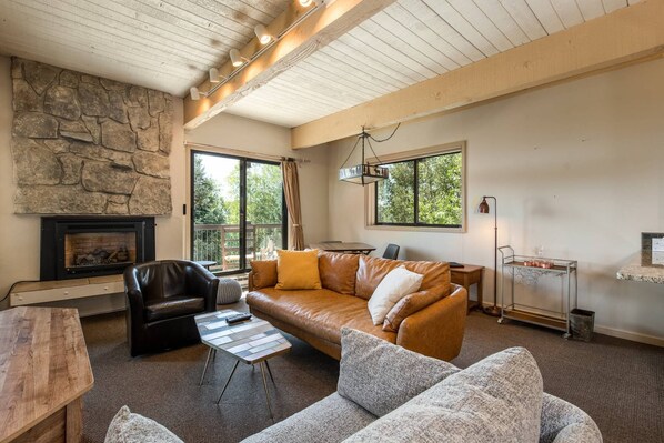 Una sala de estar acogedora con pared de piedra, chimenea, sofá de cuero, mesa de centro y un gran ventanal con vistas a la vegetación.