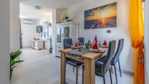 Een eetruimte met een houten tafel voor zes personen, omringd door grijs gestoffeerde stoelen. Achtergrond zichtbaar is een wit kastje met glazen deuren, en aan de muur hangt een schilderij van een zonsondergang.