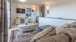 Een moderne woonkamer met een witte bank, een salontafel en een aan de muur bevestigde televisie. Op de vloer ligt een tapijt met een patroon en er is een raam met gordijnen.