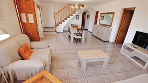 Amplia sala de estar con sofá, mesa de centro, mesa de comedor con sillas, una escalera y un mueble para la televisión.