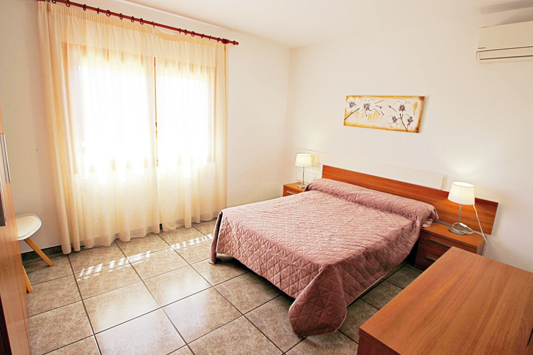 A hotel room with a large bed, two bedside tables with lamps, a small table, a chair, and a window with curtains.