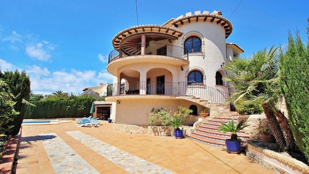 Una casa de dos pisos con un balcón circular, una piscina y una escalera de piedra que lleva a la entrada.