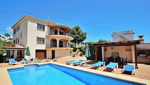 Un área de piscina con sillones de descanso y sombrillas, un edificio de dos pisos con balcones y una zona de barbacoa.