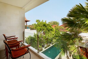 A balcony with wooden chairs, a swimming pool, and a view of a red-roofed building surrounded by greenery.