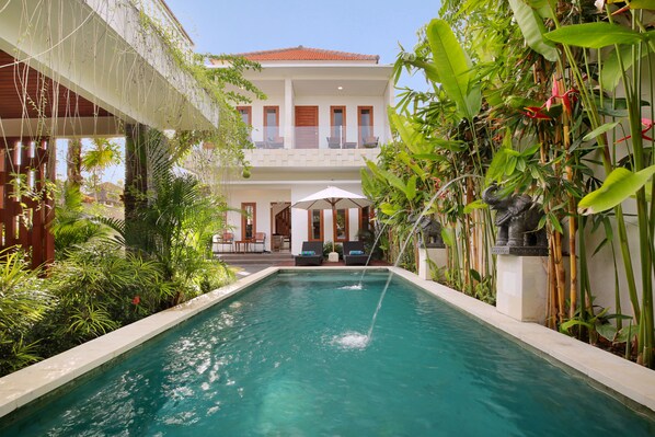 A swimming pool with a view of a building with balconies and a covered area with chairs and umbrellas.