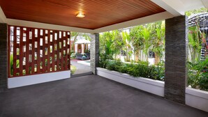 A covered outdoor area with a wooden ceiling, a white wall, and a view of a garden with plants and a statue. - Askara Villa Uluwatu (Ungasan)