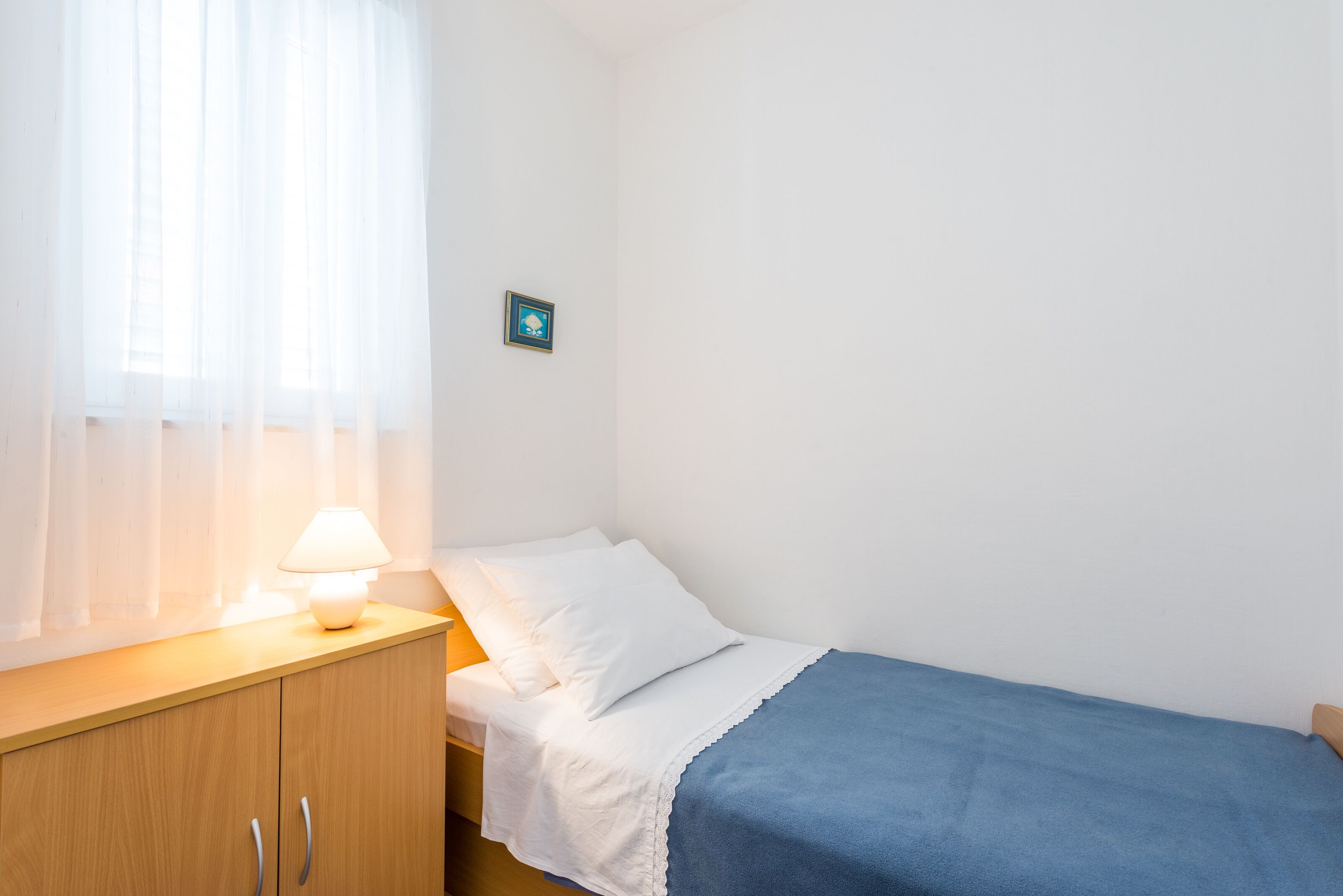 A small, neatly made bedroom with a single bed, a wooden nightstand with a lamp, and a small picture on the wall.