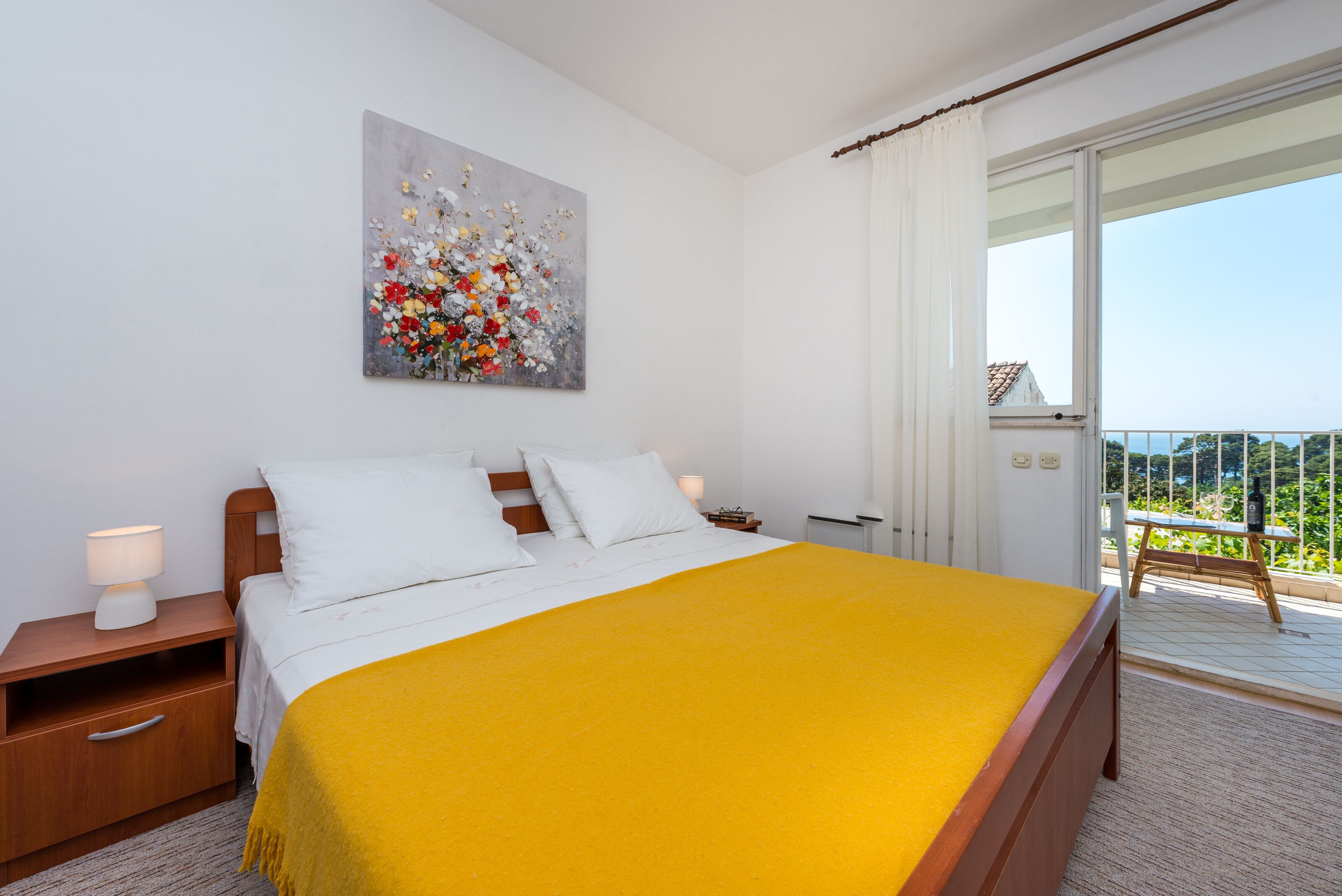 A bedroom with a bed, a nightstand, a lamp, a painting, and a balcony with a view.