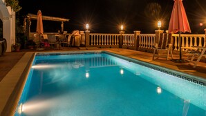 A swimming pool with a tiled edge, surrounded by lounge chairs and umbrellas, and a pergola with lights. - Casa Biontica (Cómpeta)