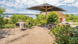 A patio area with a table and chairs, a large umbrella, and a view of a lake and thatched-roof building. - Lovely home in Mariager with WiFi (Mariager)