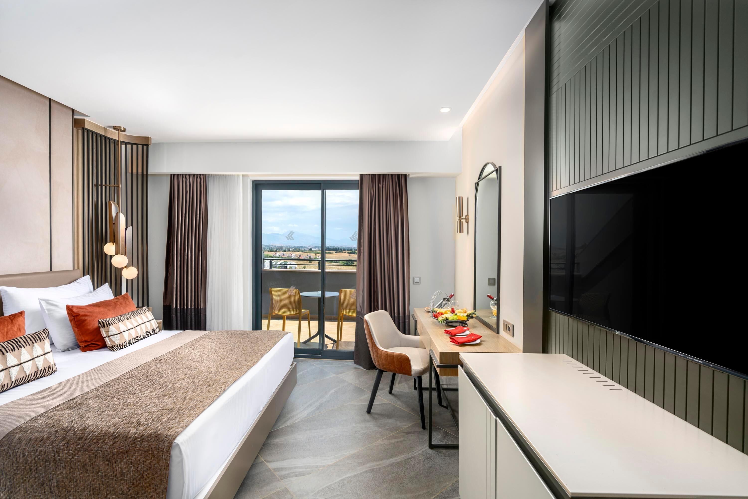 A modern hotel room with a large bed, a desk, a chair, a TV, and a balcony with a city view.