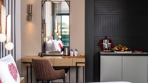Ein Hotelzimmer mit Holzschreibtisch, einem Sessel, einem Bett, einem großen Spiegel und einer Küchenzeile mit Obstschale und Wasserkocher.