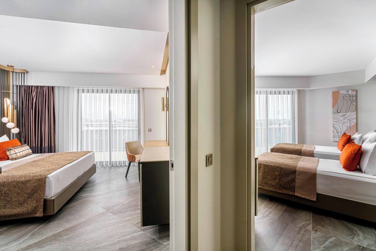 A hotel room with a bed, a desk, and a chair. There is a window with curtains and a painting on the wall.