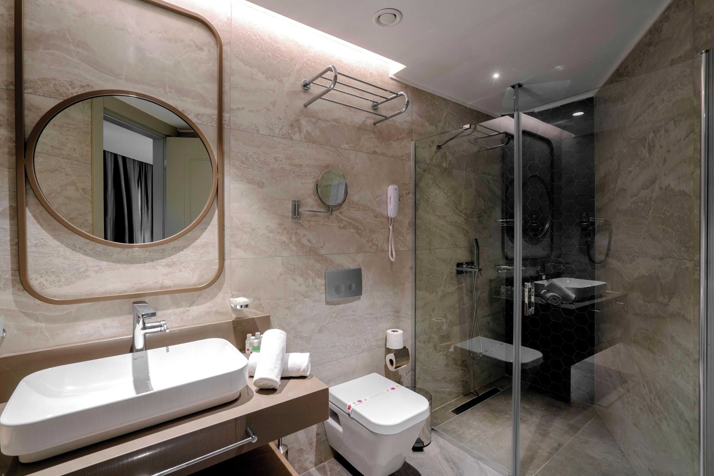 Une salle de bain moderne avec un grand miroir ovale, un lavabo rectangulaire, des toilettes et une douche séparée par une paroi de verre.