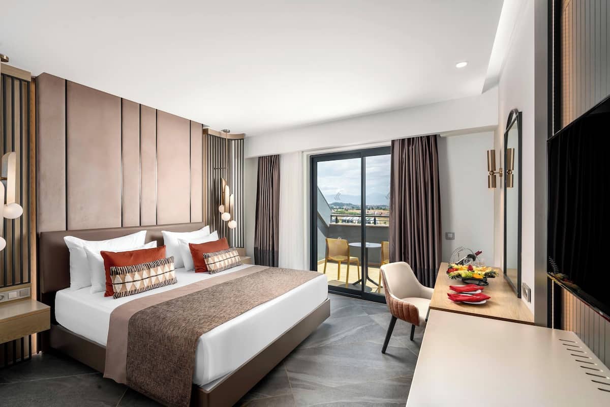A modern hotel room with a large bed, a desk with a flat-screen TV, and a balcony with a view.