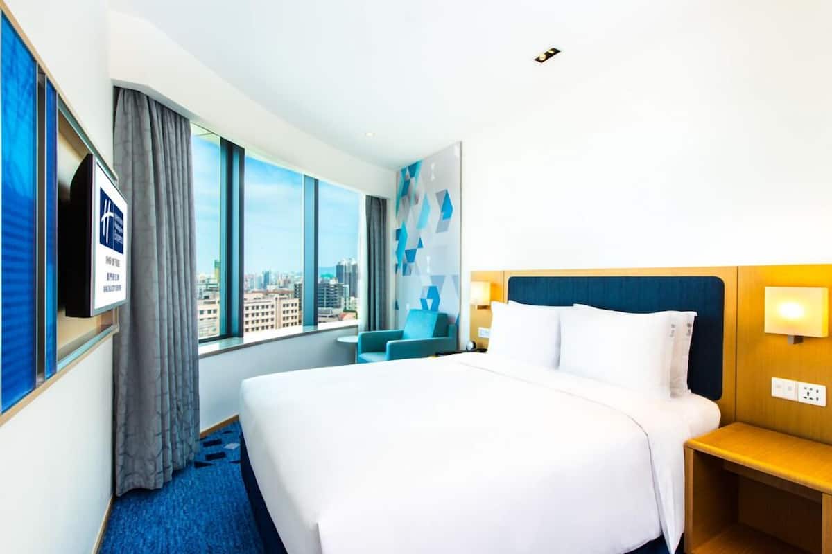 A hotel room with a large bed, a TV mounted on the wall, a window with a city view, and a blue carpet.