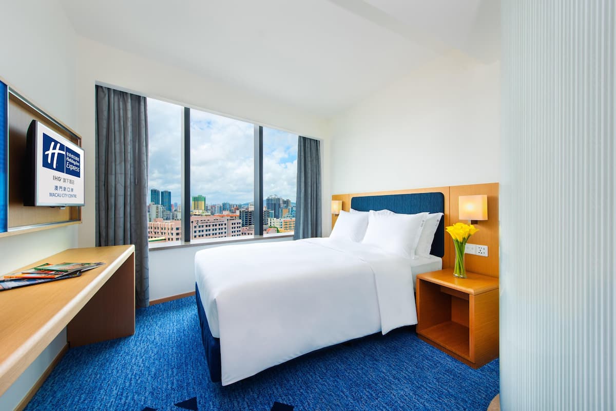 A hotel room with a bed, a desk, a TV, and a view of the city.