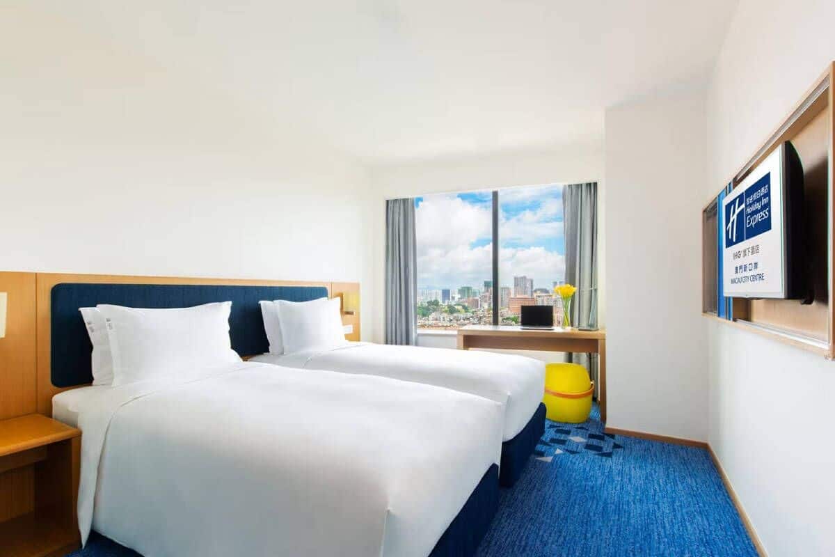 A hotel room with two beds, a desk, a chair, a window with a city view, and a sign indicating the hotel's location.