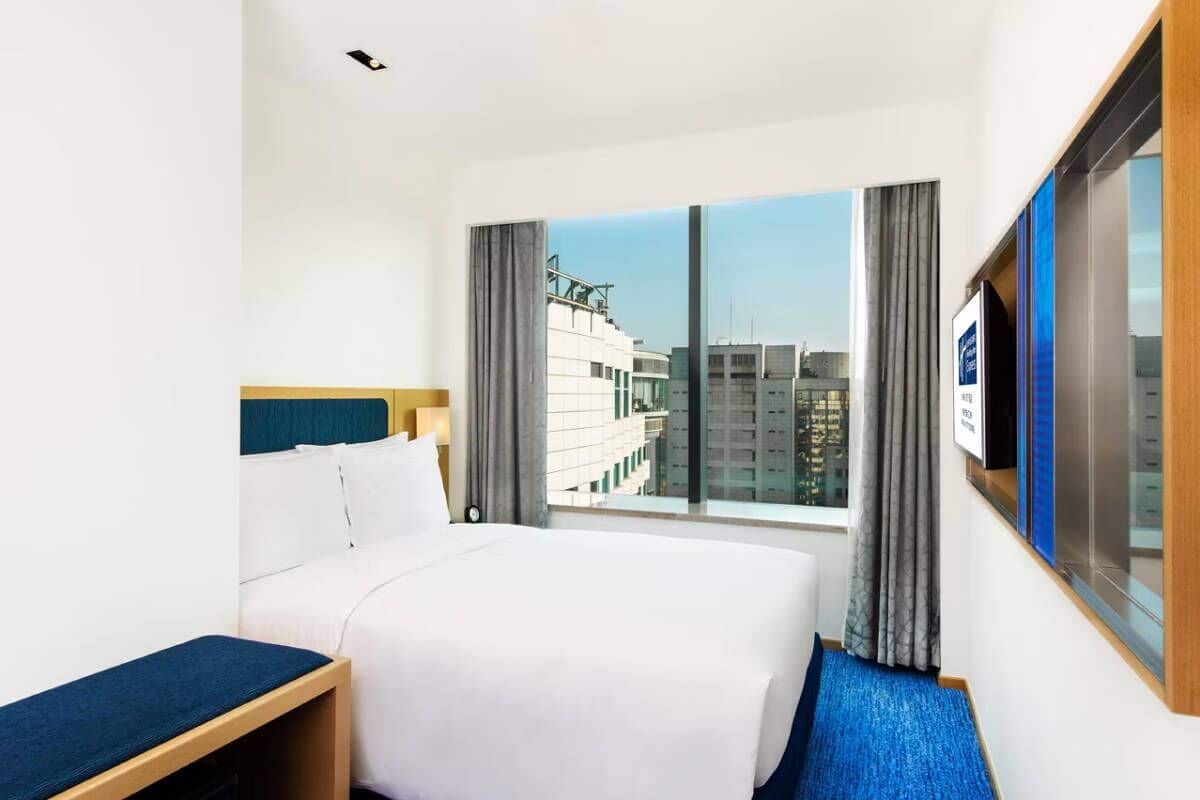 A hotel room with a bed, a desk, and a large window offering a city view.