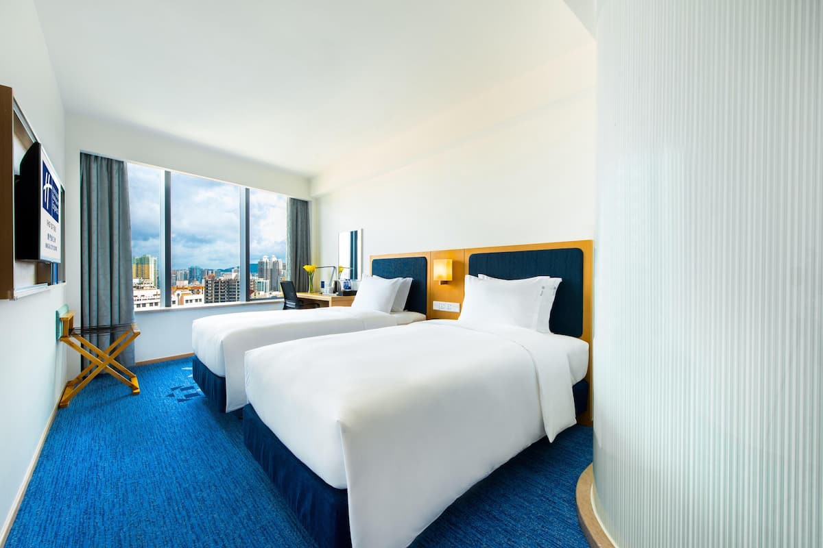 A hotel room with two beds, a city view, a blue carpet, and a wooden side table.
