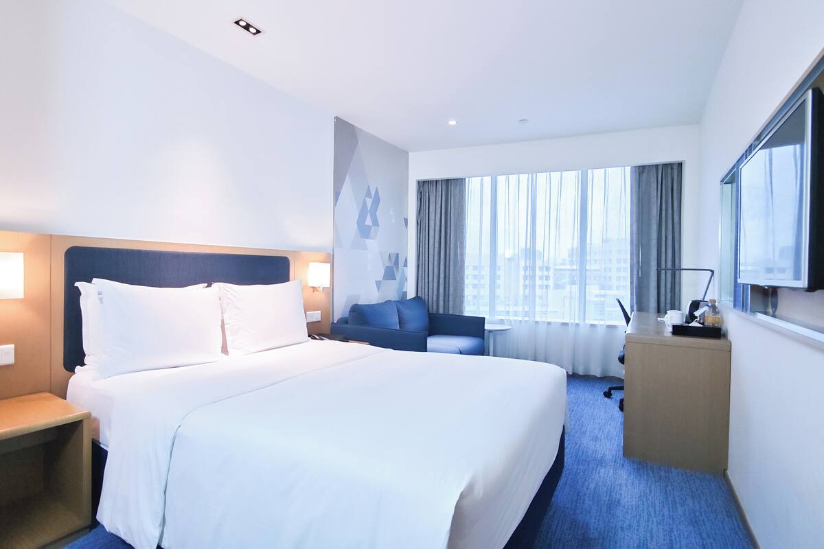 A hotel room with a large bed, a desk, a chair, a TV, and a window with curtains.