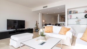 Een moderne woonkamer met een flatscreen televisie, witte banken, een salontafel en een boekenkast.