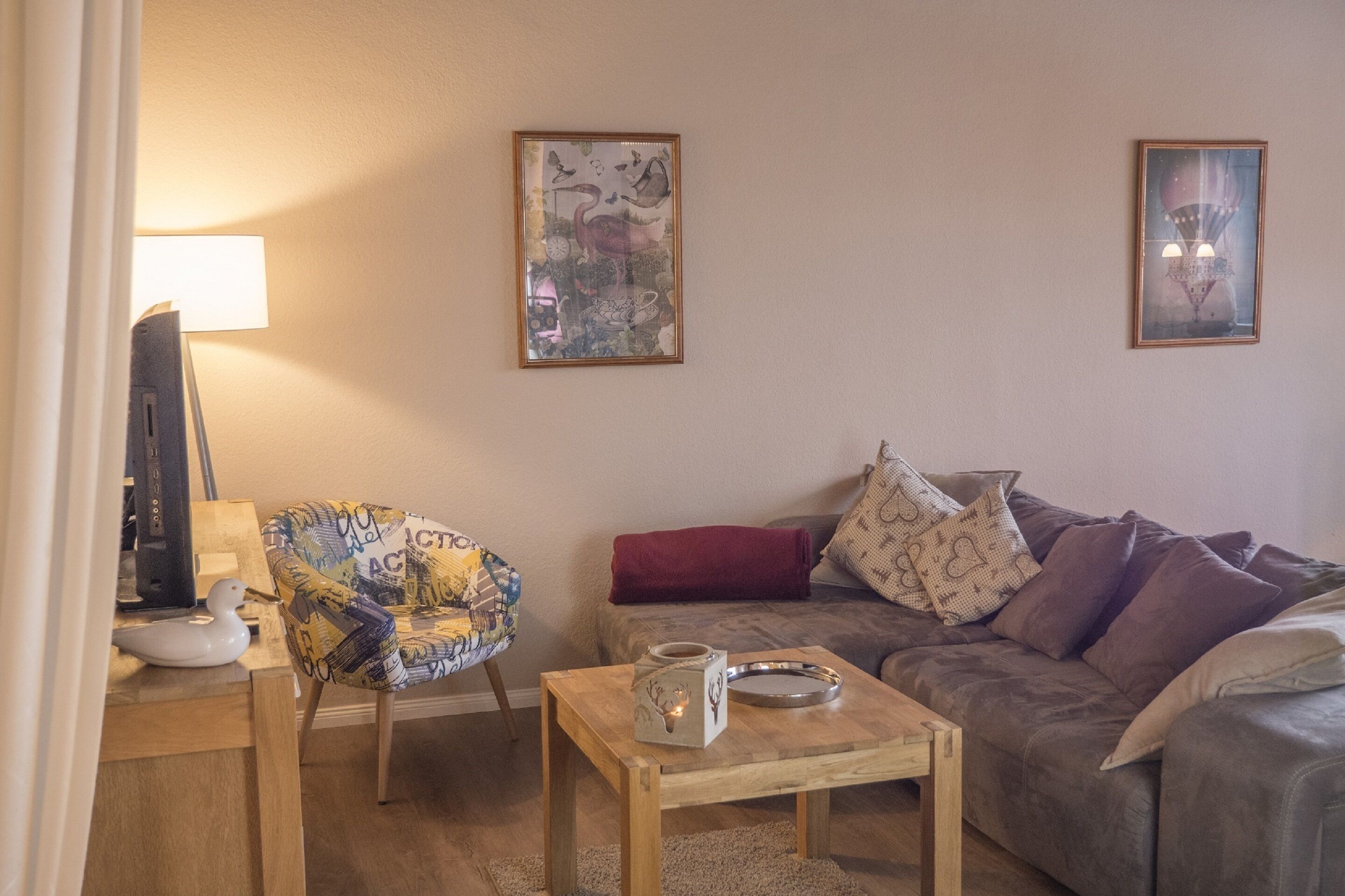 A living room with a wooden coffee table, a patterned armchair, a grey sofa with cushions, and two framed artworks on the wall.