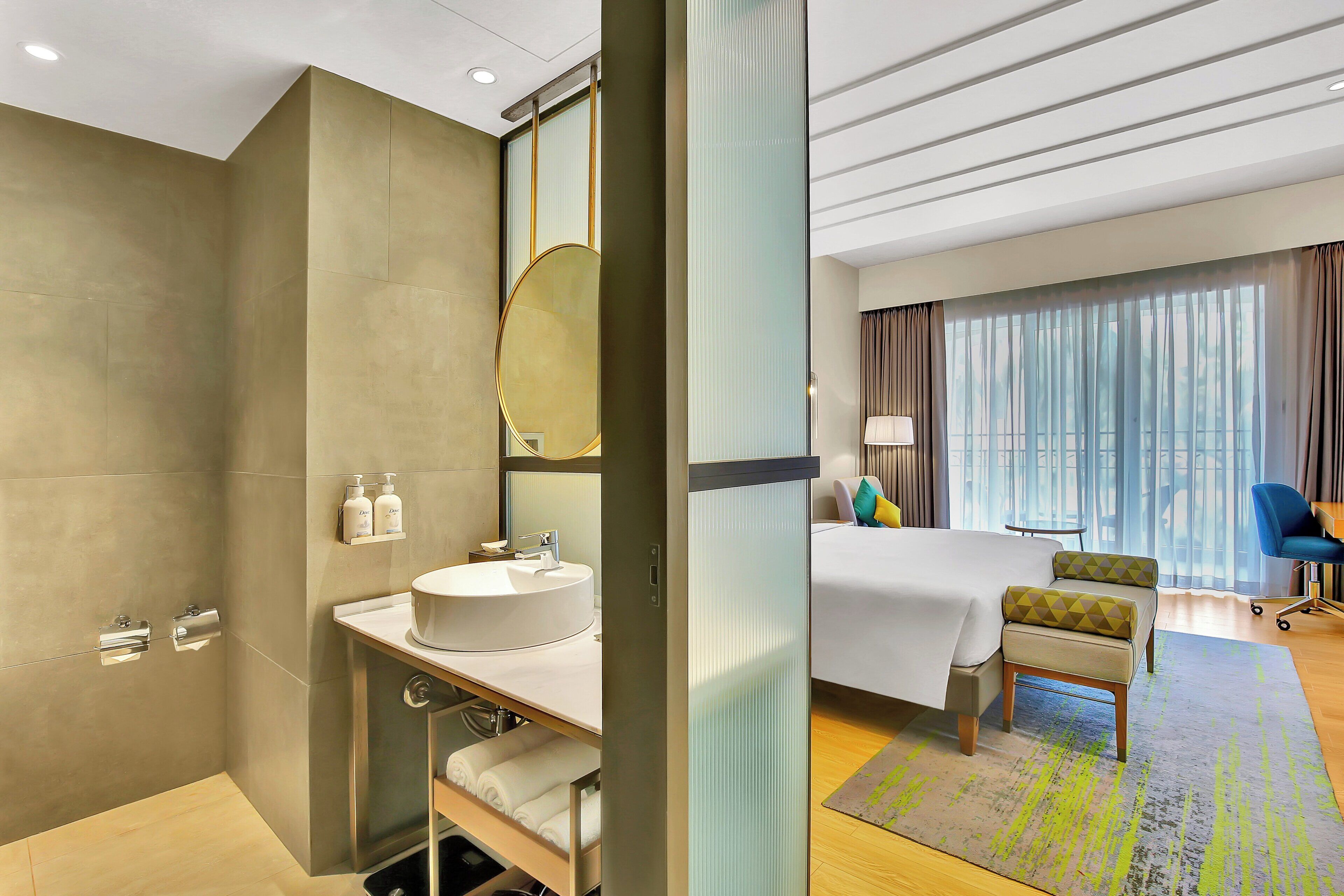 Een moderne hotelkamer met een groot bed, een zithoek met een stoel en een klein tafeltje, een badkamer met een wastafel en spiegel, en een groot raam met gordijnen.