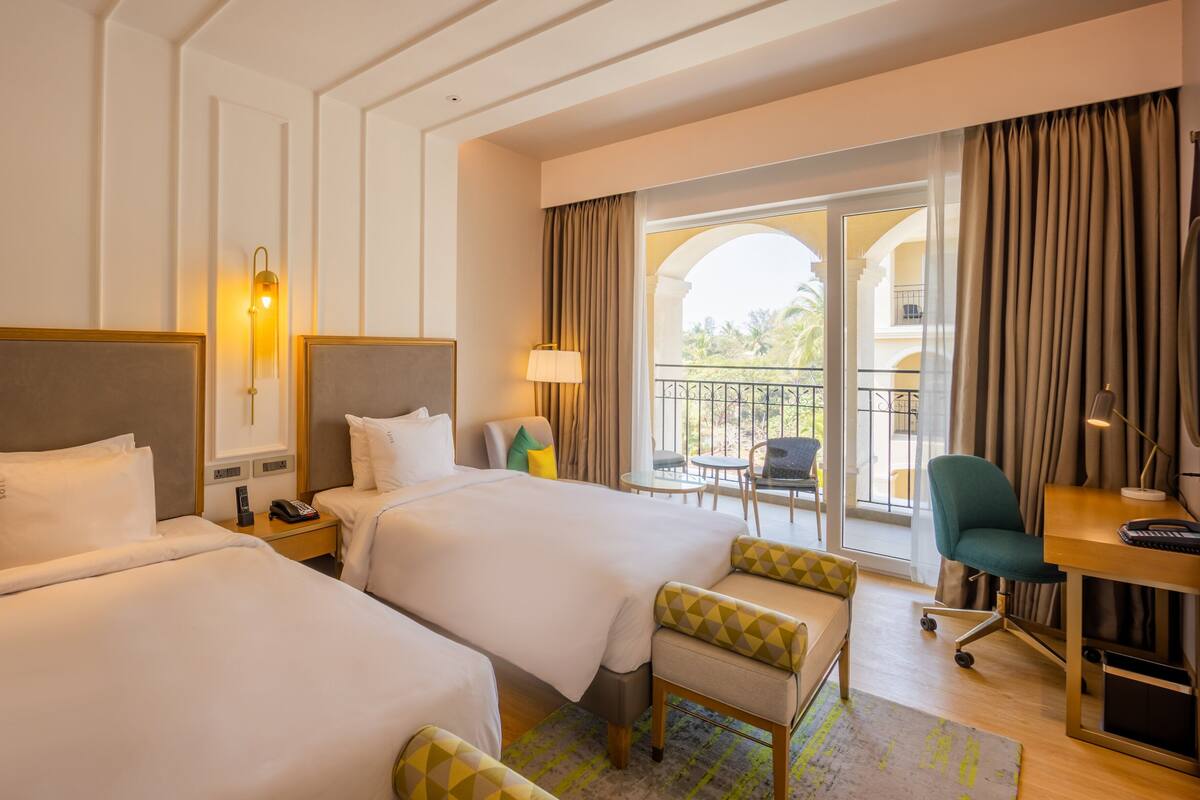 A hotel room with two beds, a desk, a chair, and a balcony with curtains.