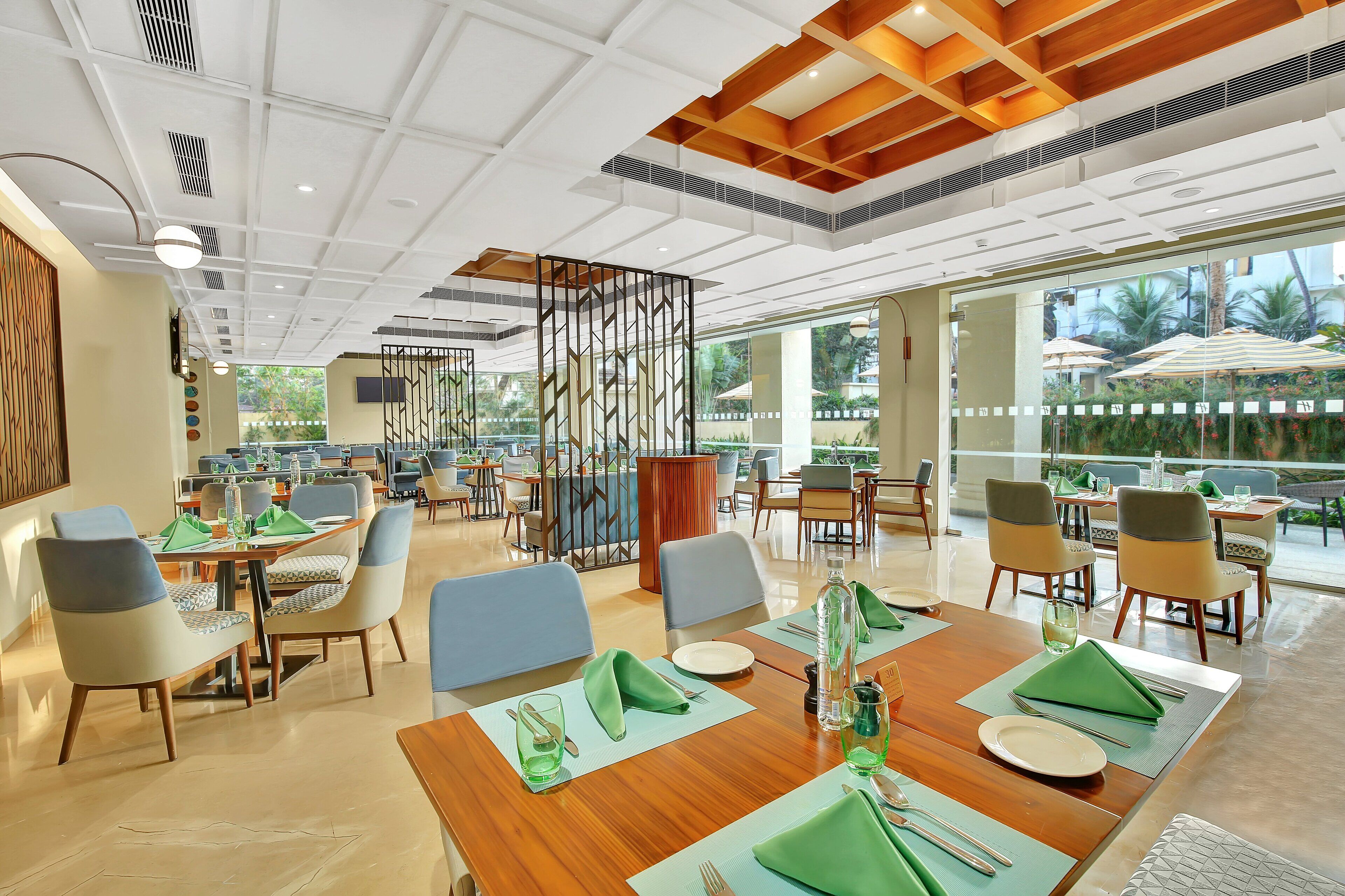Un espace de restauration moderne avec des tables et des chaises en bois, un élément de séparation décoratif et de grandes fenêtres offrant une vue sur l’extérieur.