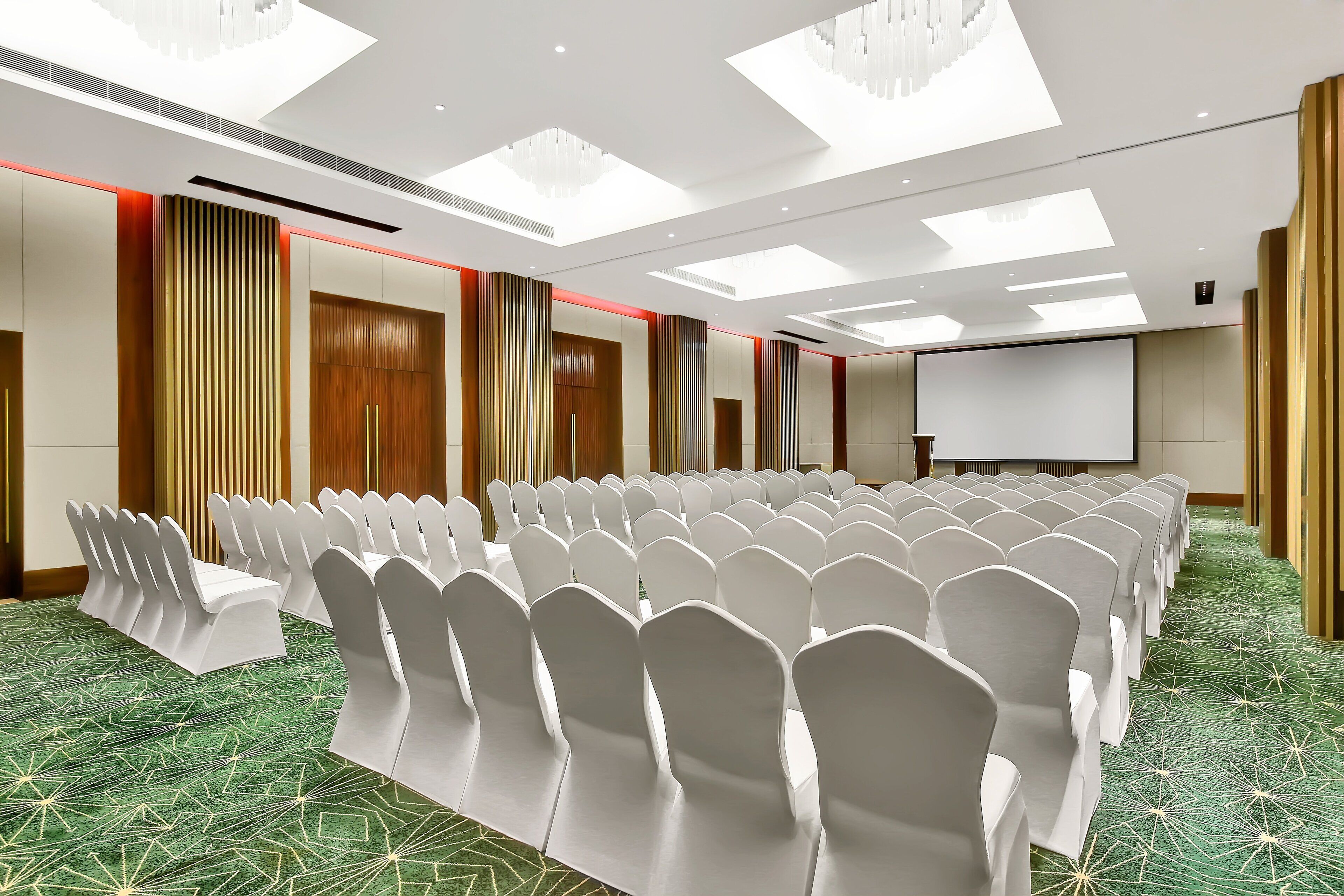 A conference room with rows of white chairs, a projector screen, and a green patterned carpet.