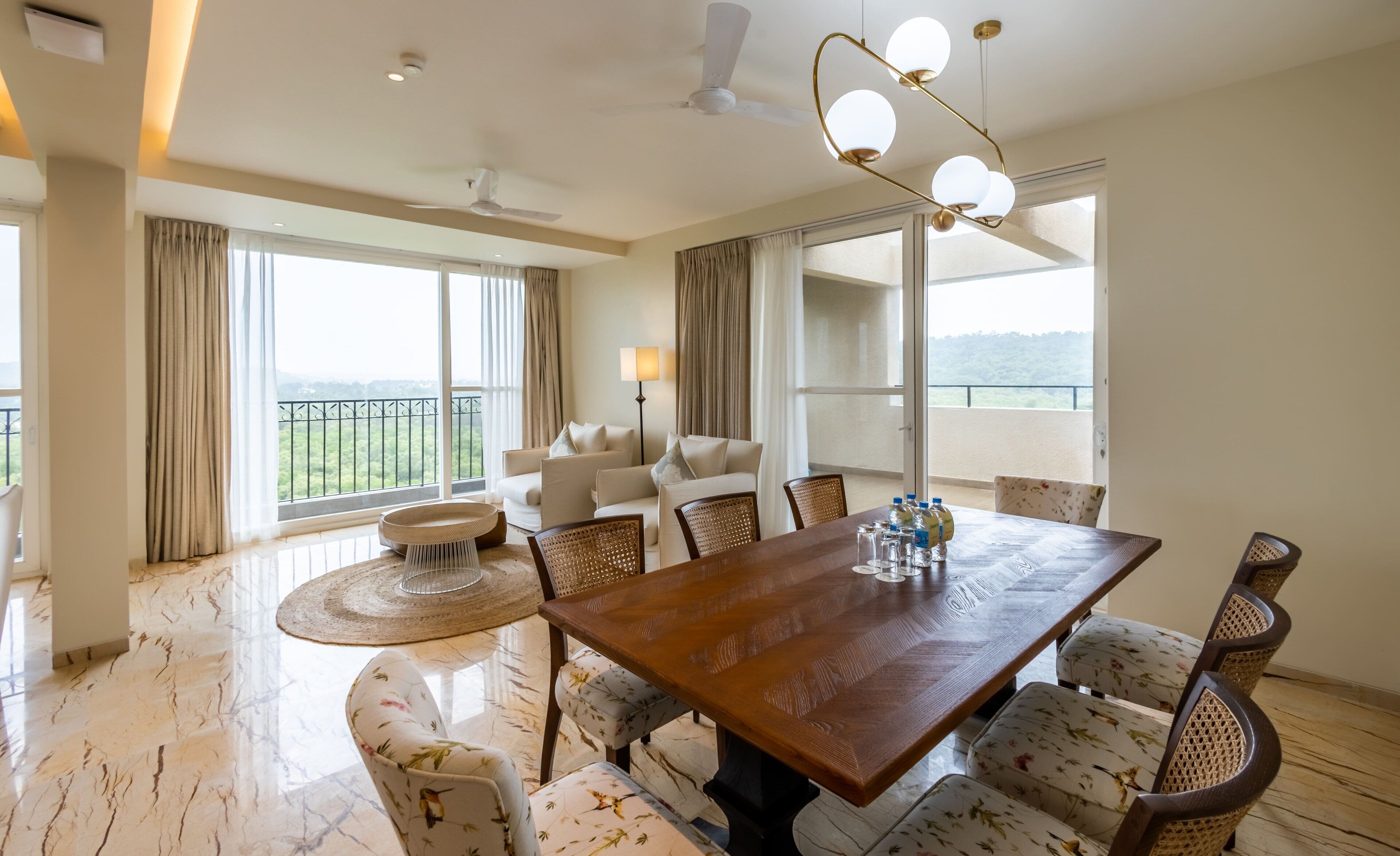 A spacious room with a dining table, chairs, and a sofa. There are large windows with curtains, a ceiling fan, and a lamp.
